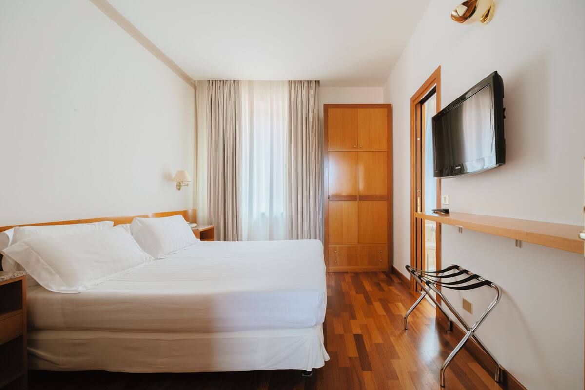 A hotel room with a bed, a TV mounted on the wall, a wooden wardrobe, and a window with curtains.