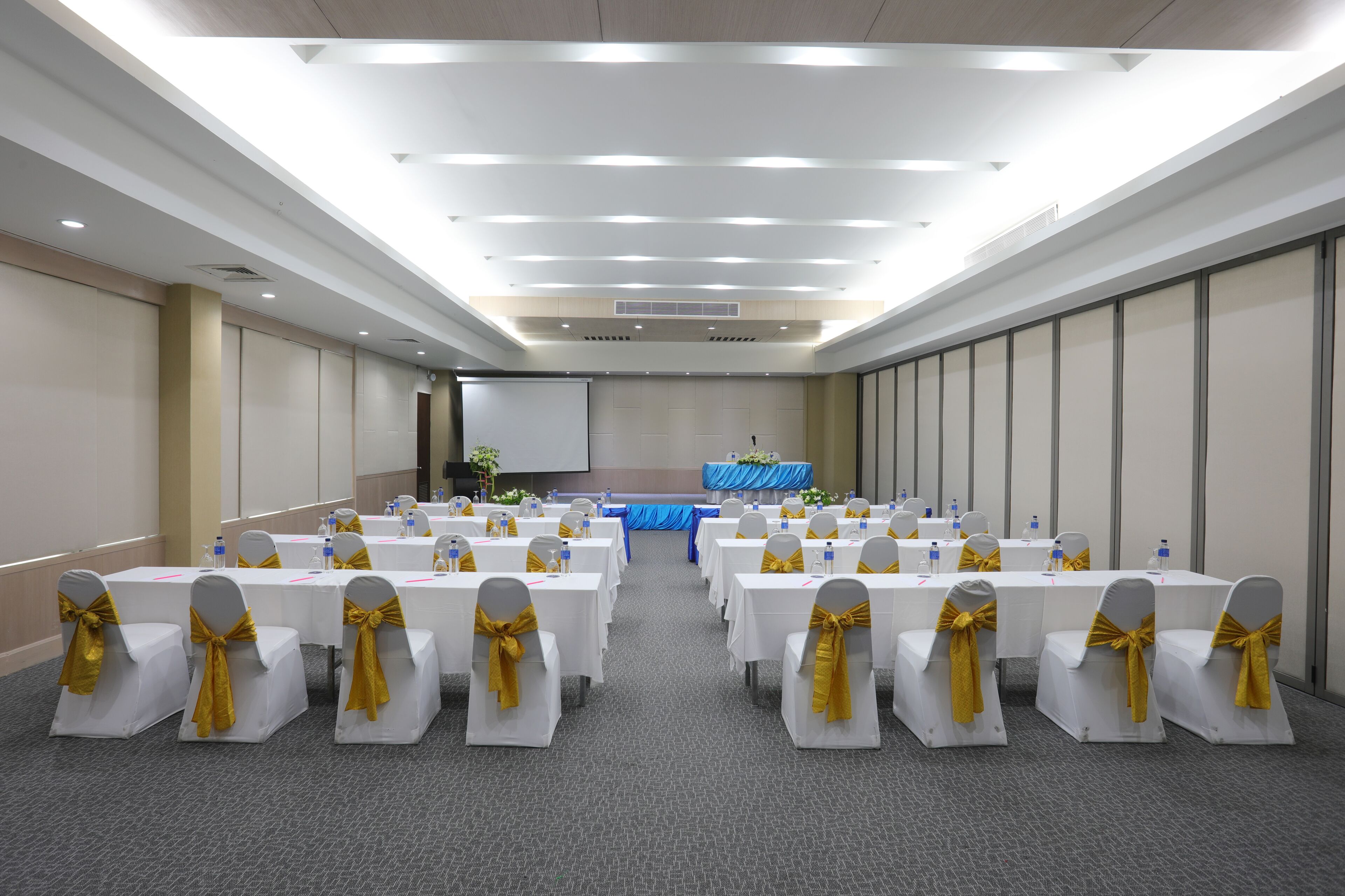 A conference room with rows of white tables and chairs, each with a gold bow.