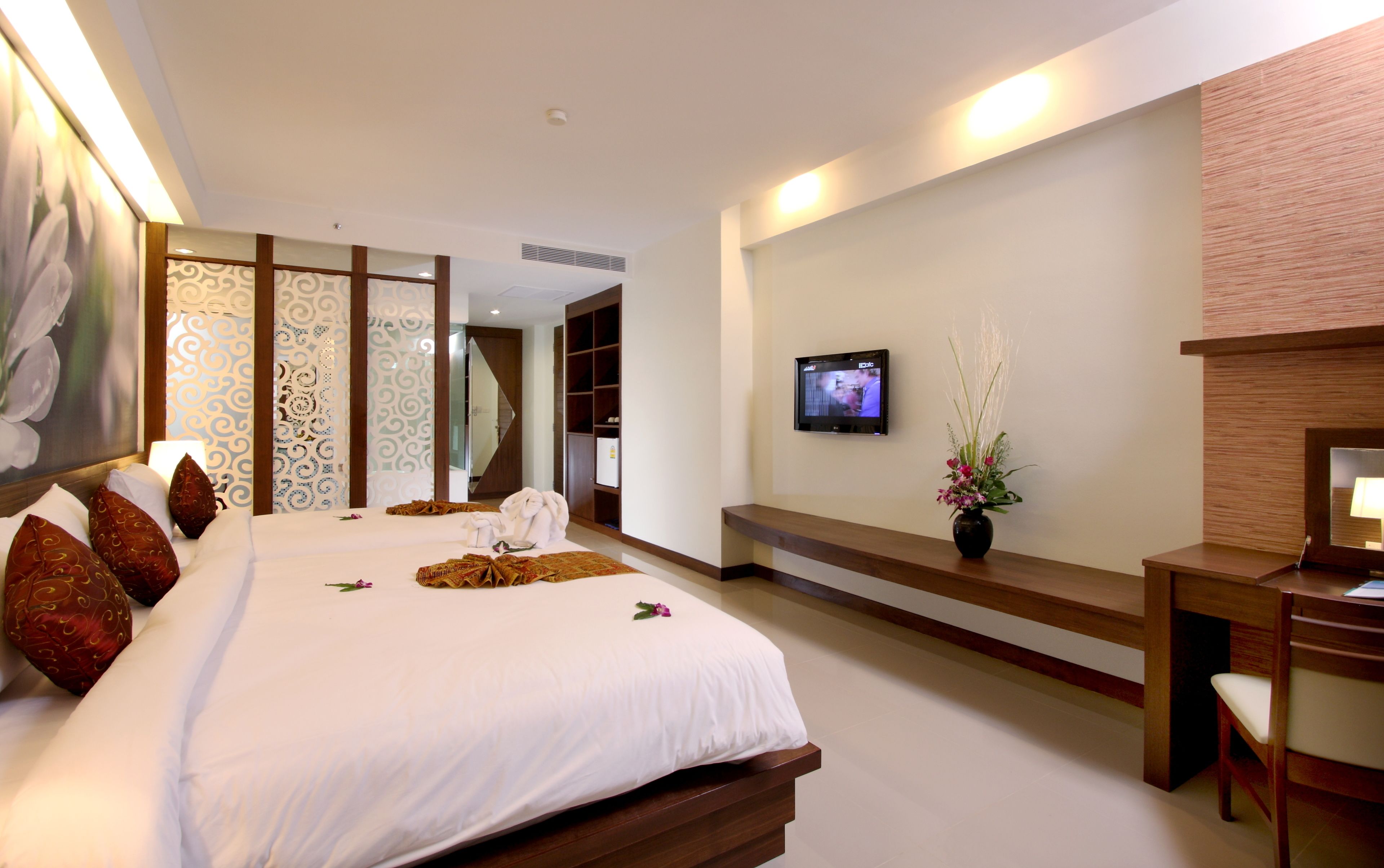 A hotel room with a large bed, a TV mounted on the wall, a desk with a chair, and a decorative vase with flowers.