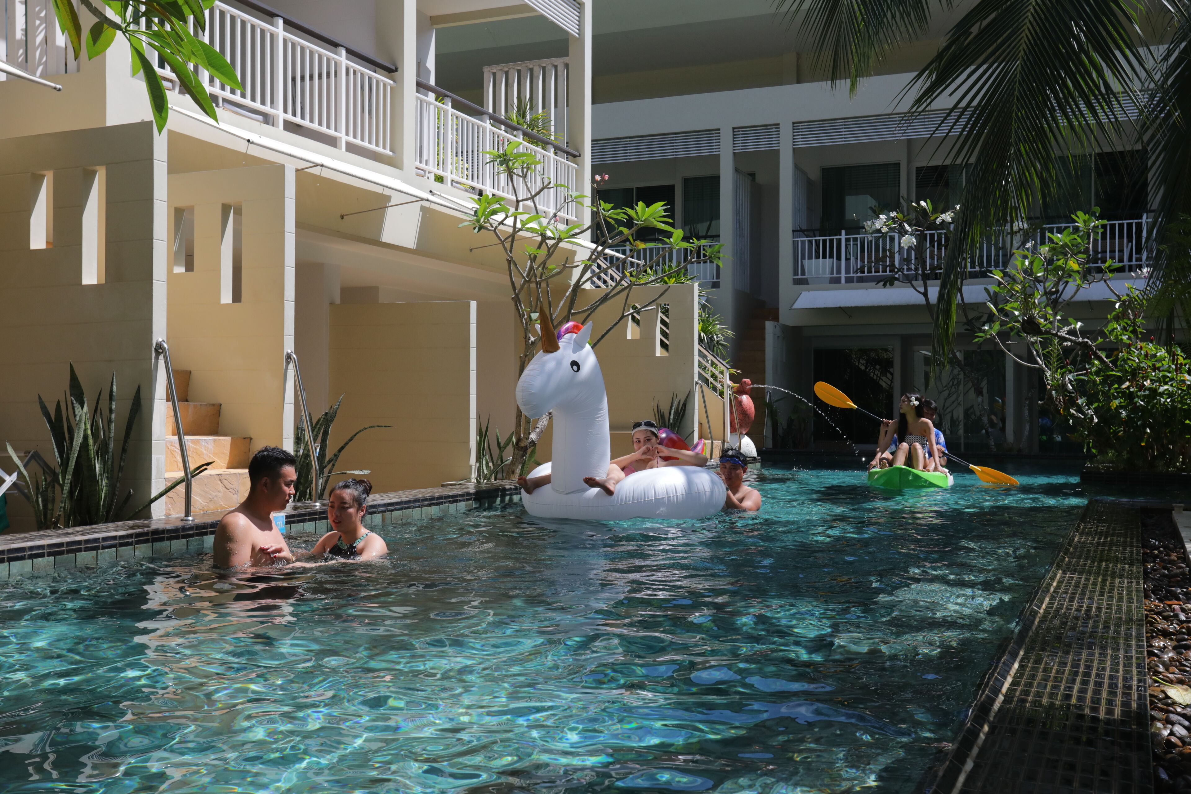 A swimming pool area with a white swan float, people in the water, and a building with balconies.