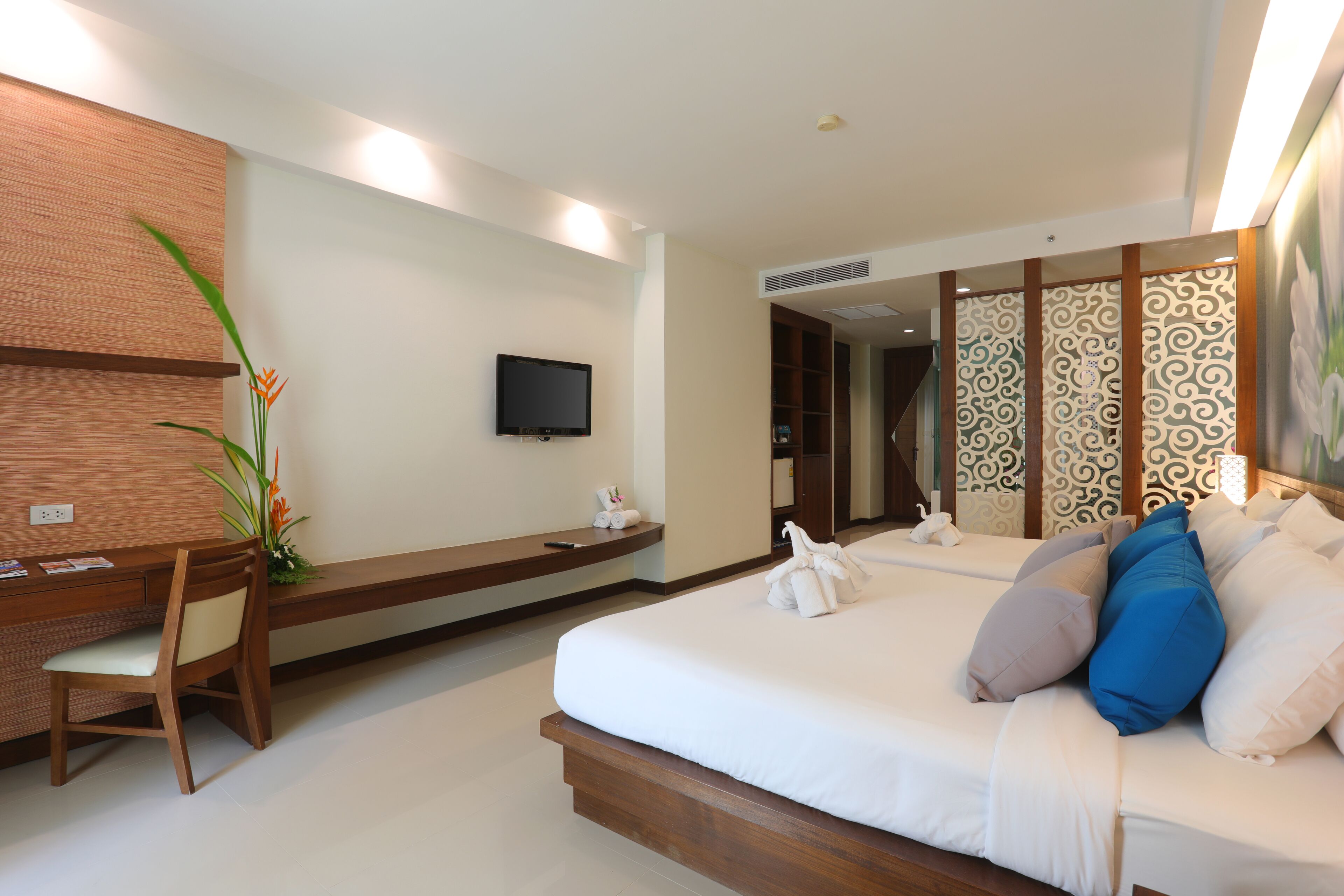 A hotel room with a wooden desk, a bed with white bedding and blue pillows, a TV on the wall, and a large plant.