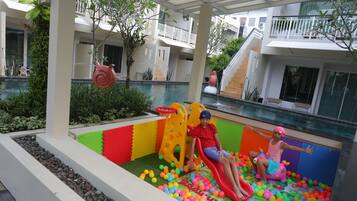 Kids can play in the vibrant play area with a colorful slide and ball pit, adjacent to a swimming pool.