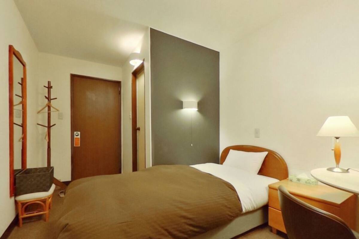 Une chambre d’hôtel avec un lit simple, une table de chevet avec une lampe, une chaise, un porte-manteau et une porte.
