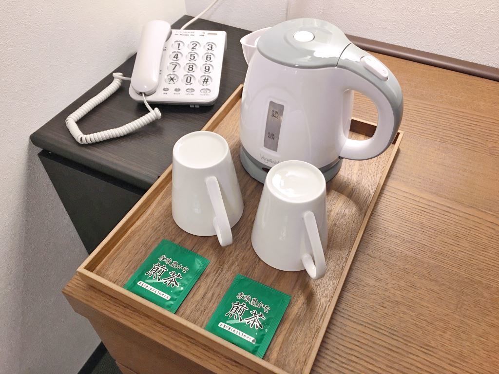 A wooden tray with two white mugs, a white electric kettle, a white telephone, and two green packets on a table.