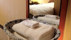 Habitación de hotel con cama, mesita de noche y espejo que refleja el espacio. En el suelo, hay dos cestas con toallas dobladas y una bolsa de plástico.