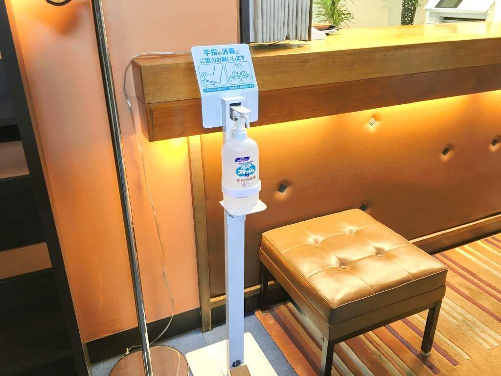A reception area with a hand sanitizer dispenser mounted on a white pole, a wooden reception desk, and a brown leather bench.