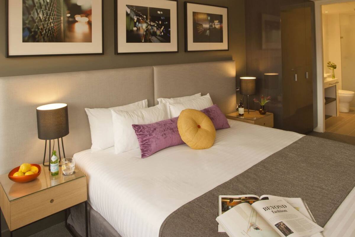 Ett hotellrum med en säng, sängbord, ett litet bord, en lampa, en tidning och inramade bilder på väggen.