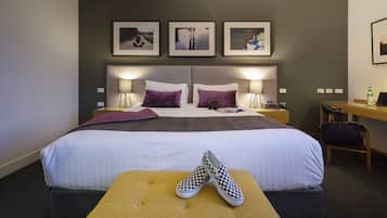 A hotel room with a large bed, two bedside tables with lamps, a desk, and a chair. There are three framed pictures on the wall above the bed.