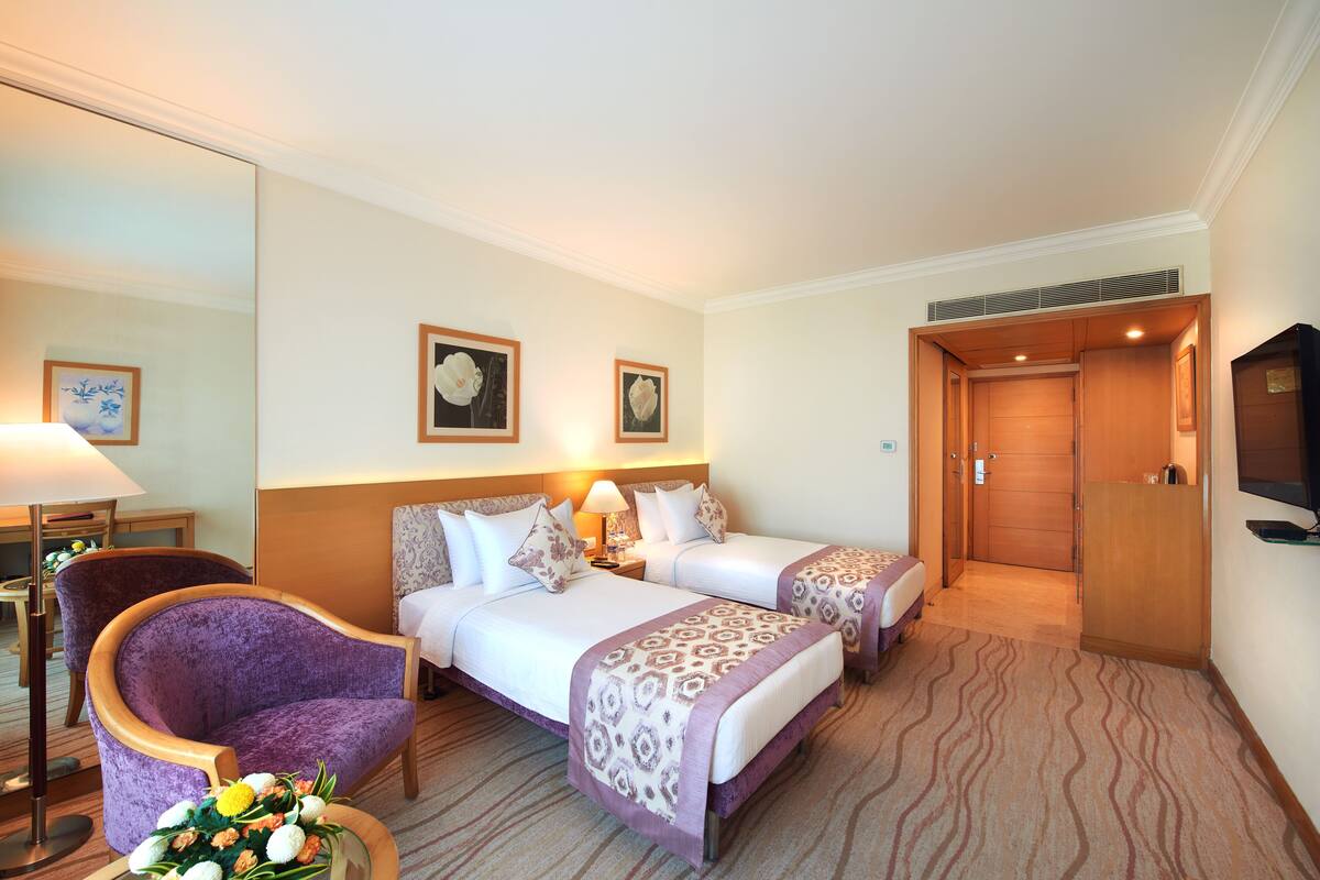 Una habitación de hotel con dos camas, un sillón morado, un escritorio con lámpara y una televisión.