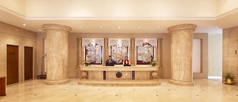 A spacious lobby with marble flooring, large columns, and a reception desk with two staff members.