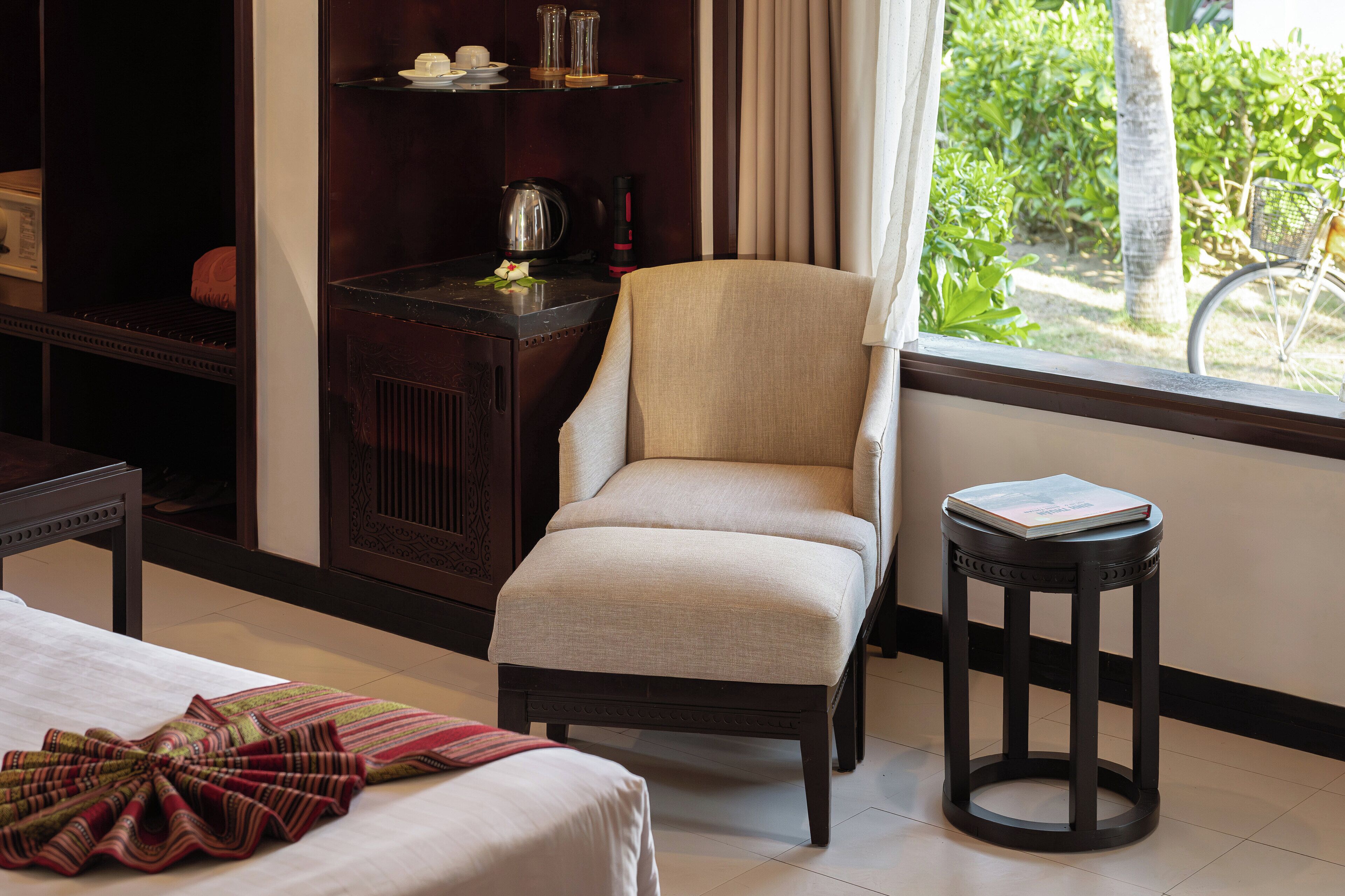 A hotel room with a bed, a chair, a small table, a shelf with items, and a view of greenery outside.