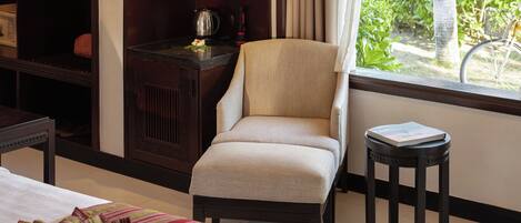A hotel room with a bed, a chair, a small table, a shelf with items, and a view of greenery outside.