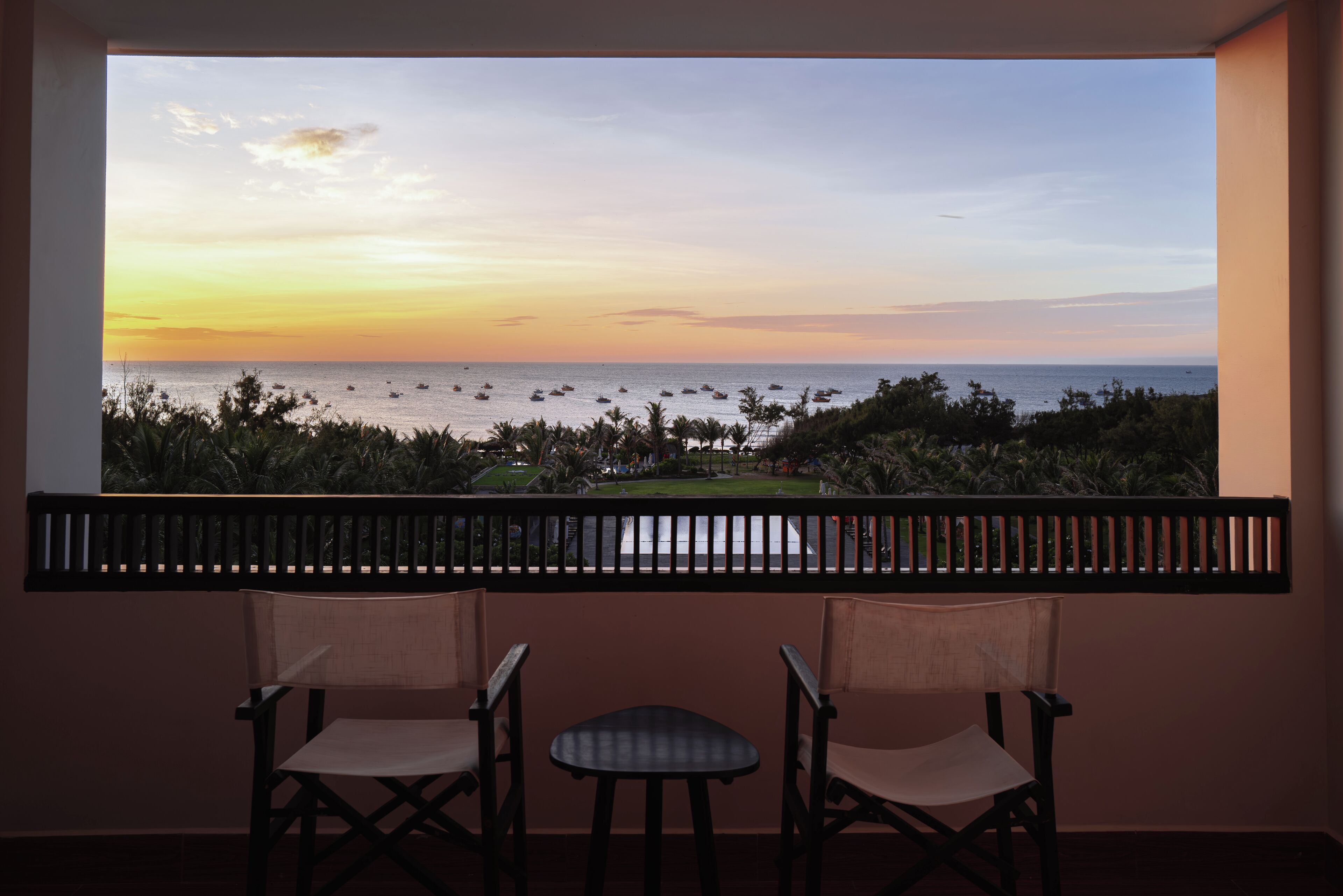 A balcony with two chairs and a small table, overlooking a beach with boats and palm trees during sunset.