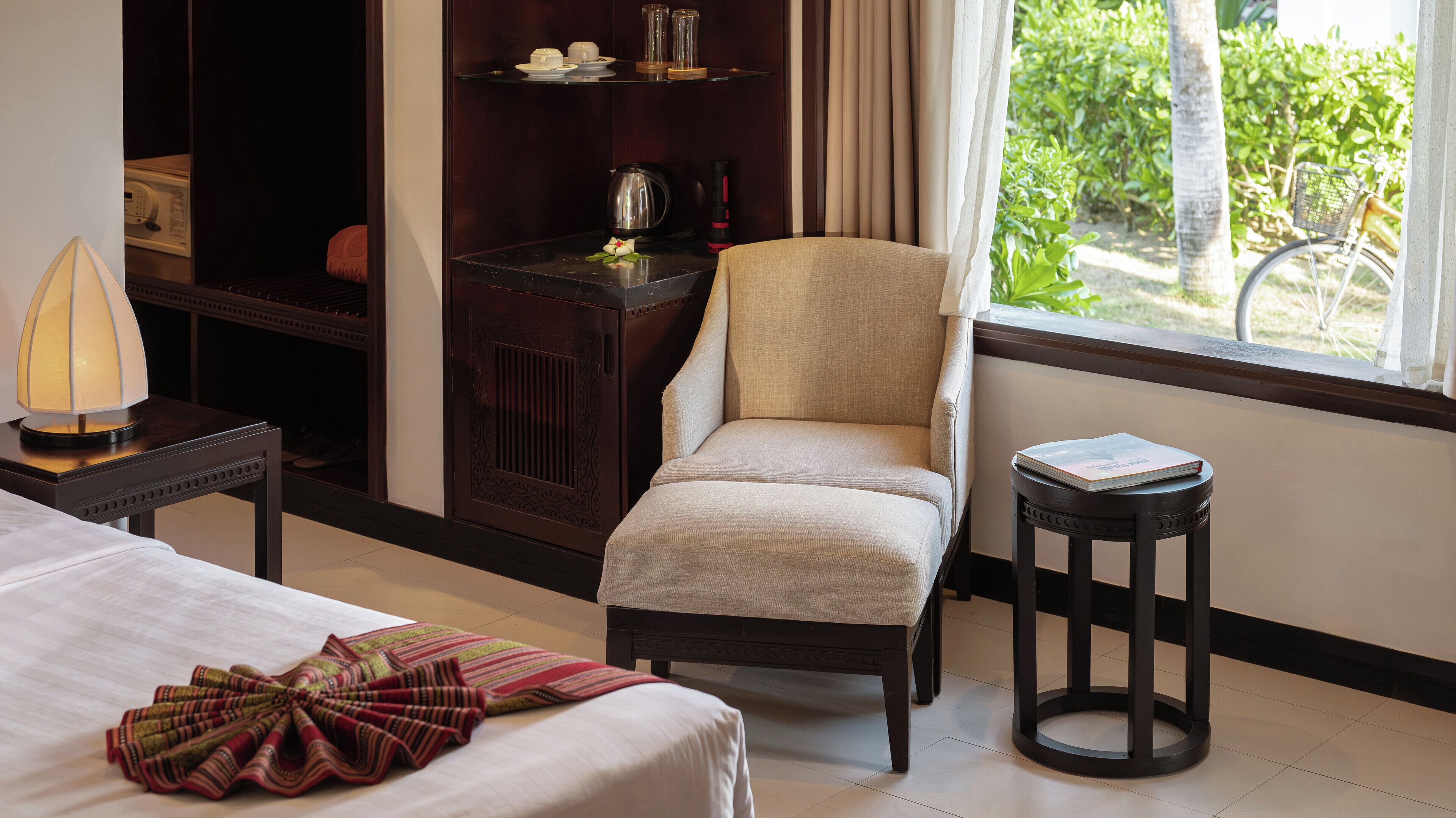 A hotel room with a bed, a chair, a small table, a bookshelf, and a window with a view of greenery.