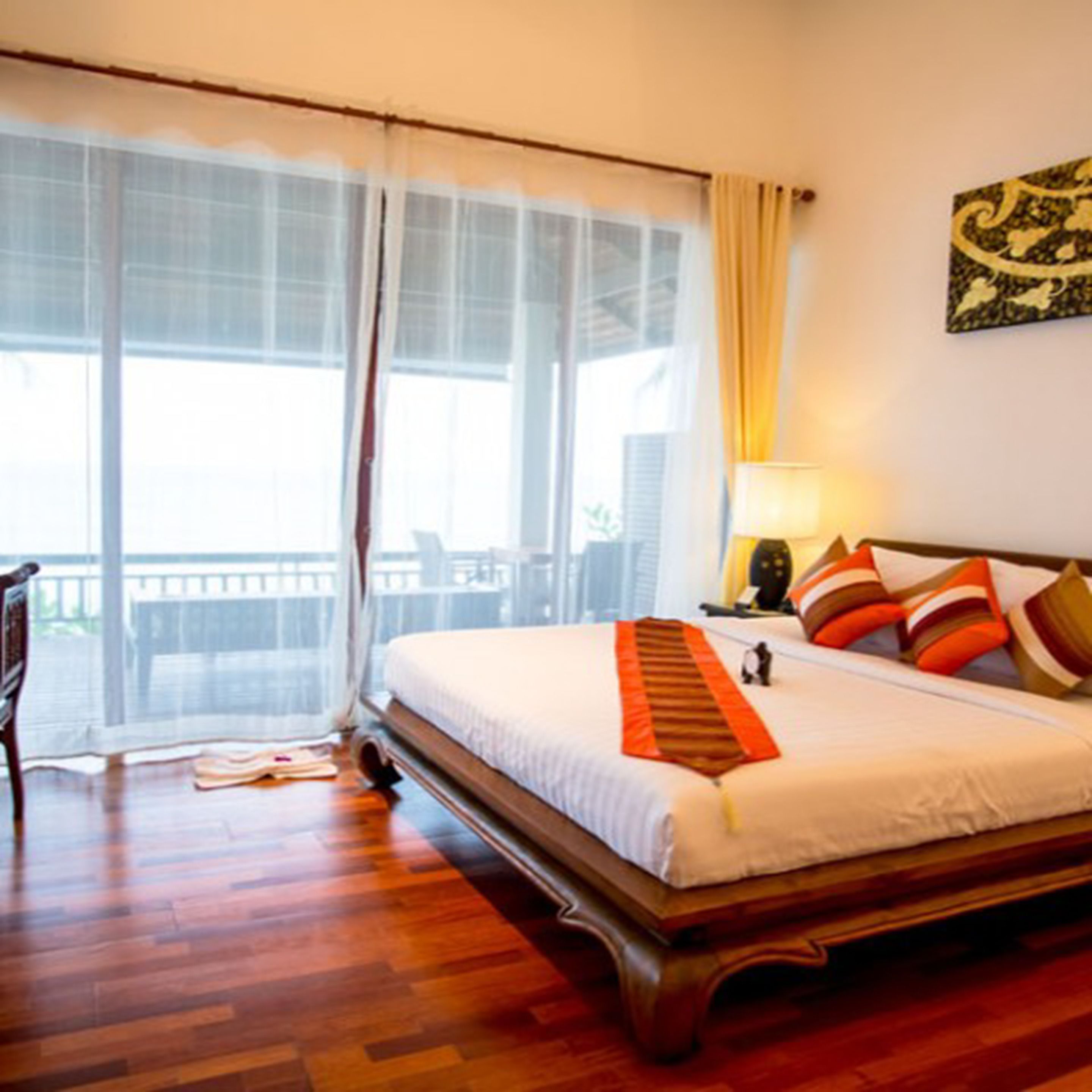 A hotel room with a large bed, a wooden floor, a balcony with a table and chairs, and a wall-mounted lamp.
