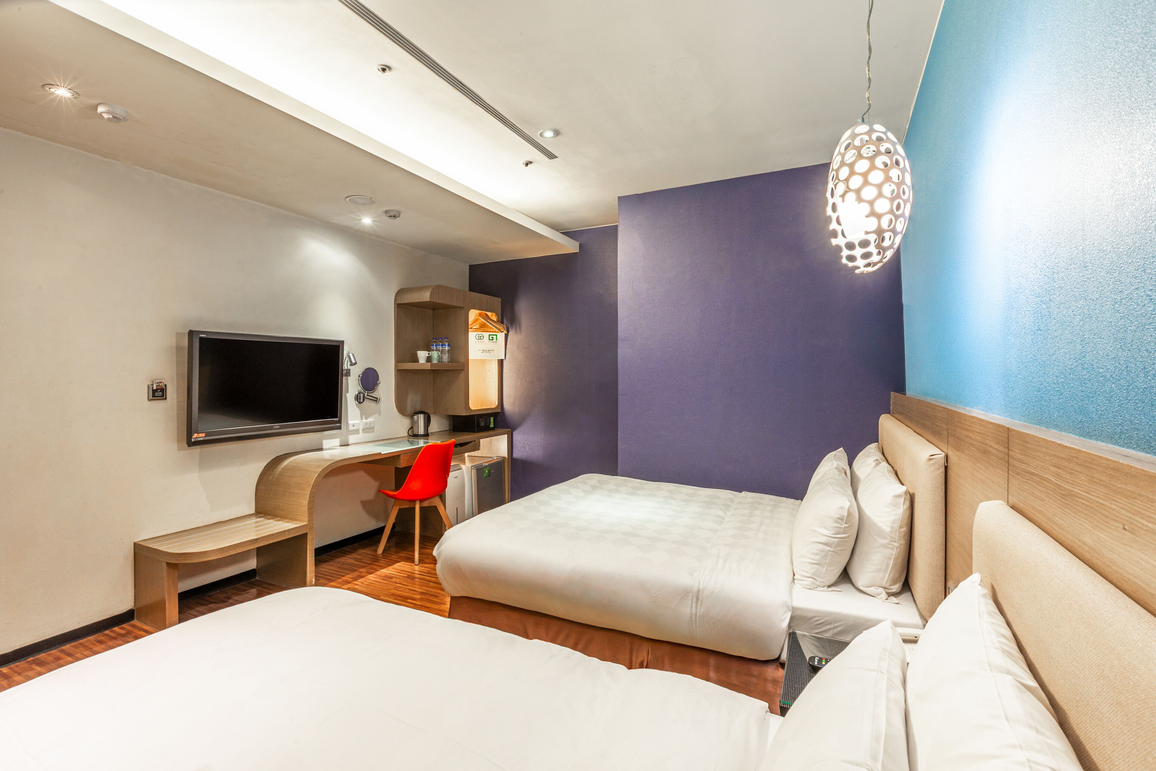 A modern hotel room with a flat-screen TV, a desk with a chair, and two beds with pillows.