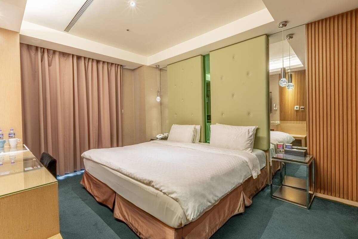 A hotel room with a large bed, a desk, a mirror, and a view of the city.
