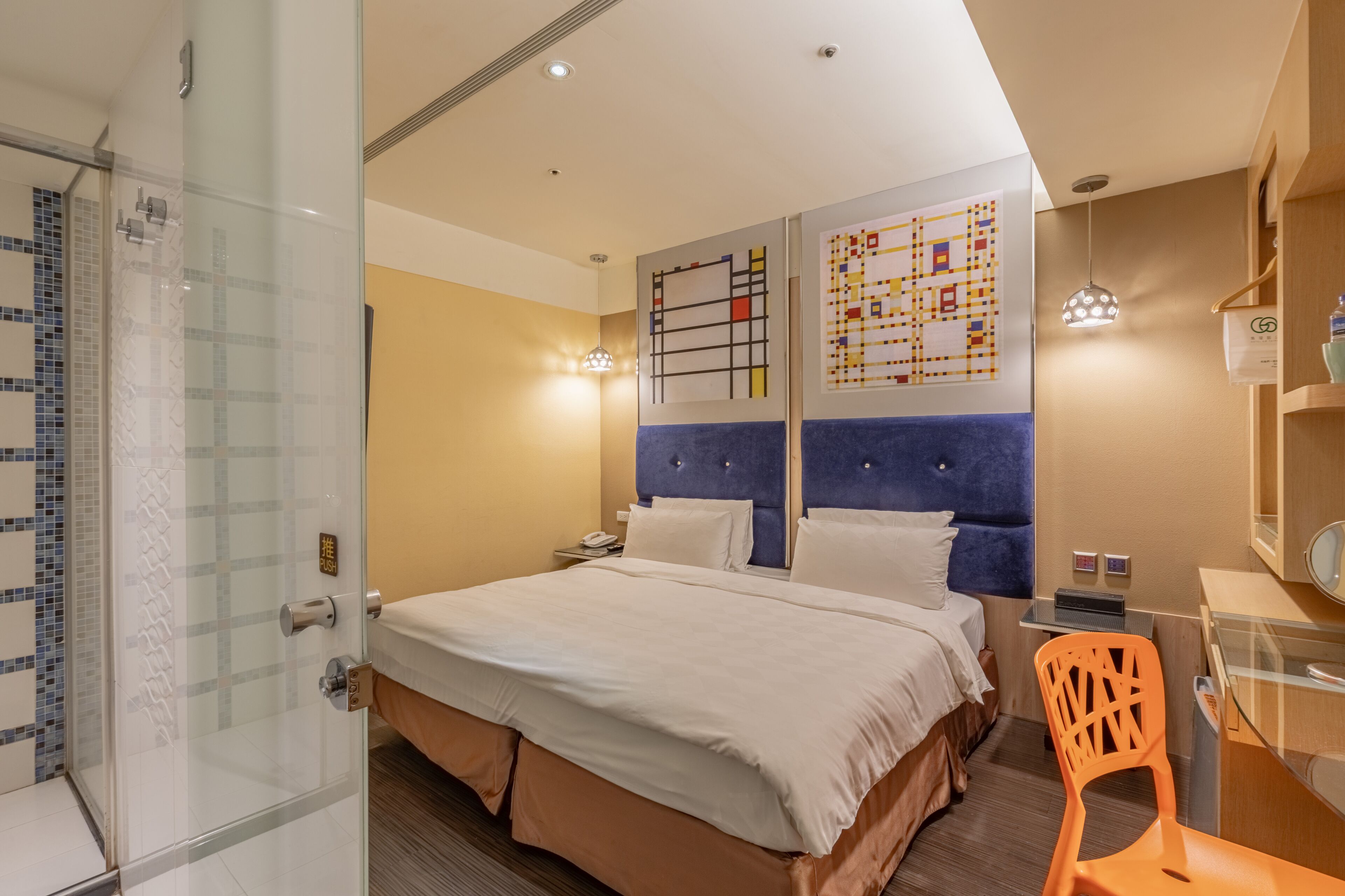 A hotel room with a glass shower enclosure, a bed with white bedding, a blue headboard, and a desk with a chair.