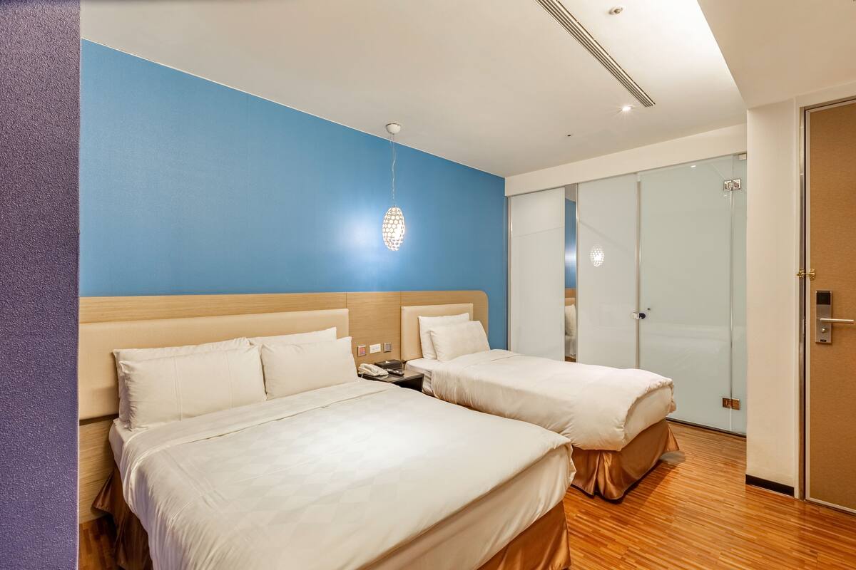 A hotel room with two beds, a blue accent wall, a wooden floor, and a glass door leading to a shower area.