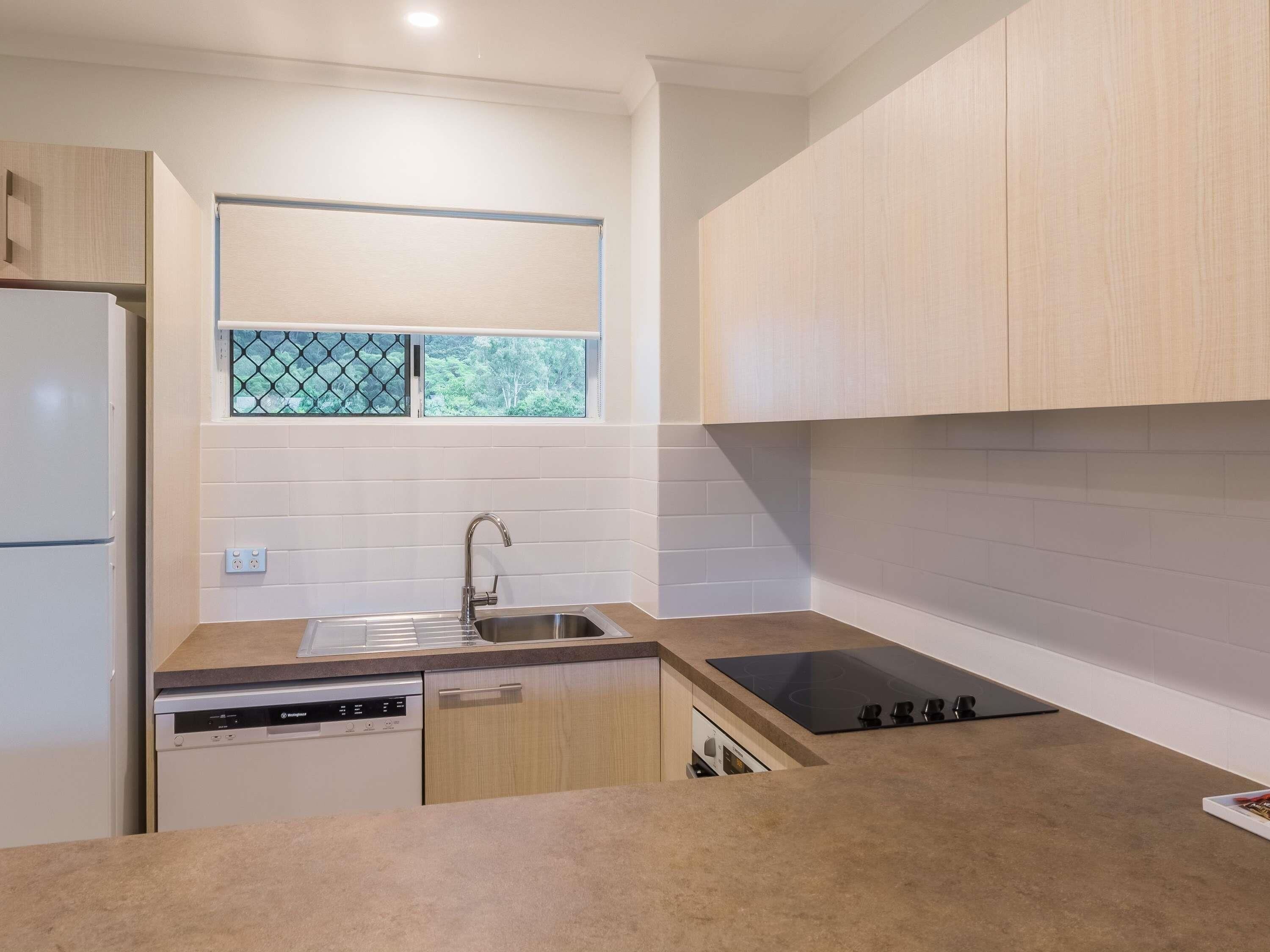 Una cocina moderna con armarios blancos, un lavamanos de acero inoxidable, lavavajillas y ventana con vista a follaje.