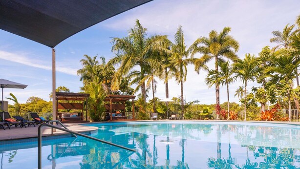 A swimming pool with lounge chairs and umbrellas, surrounded by palm trees. - The Sebel Palm Cove (Palm Cove)