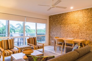 Relax in a spacious living area with a large window offering a view of palm trees, a ceiling fan, dining table, and comfortable sofa.