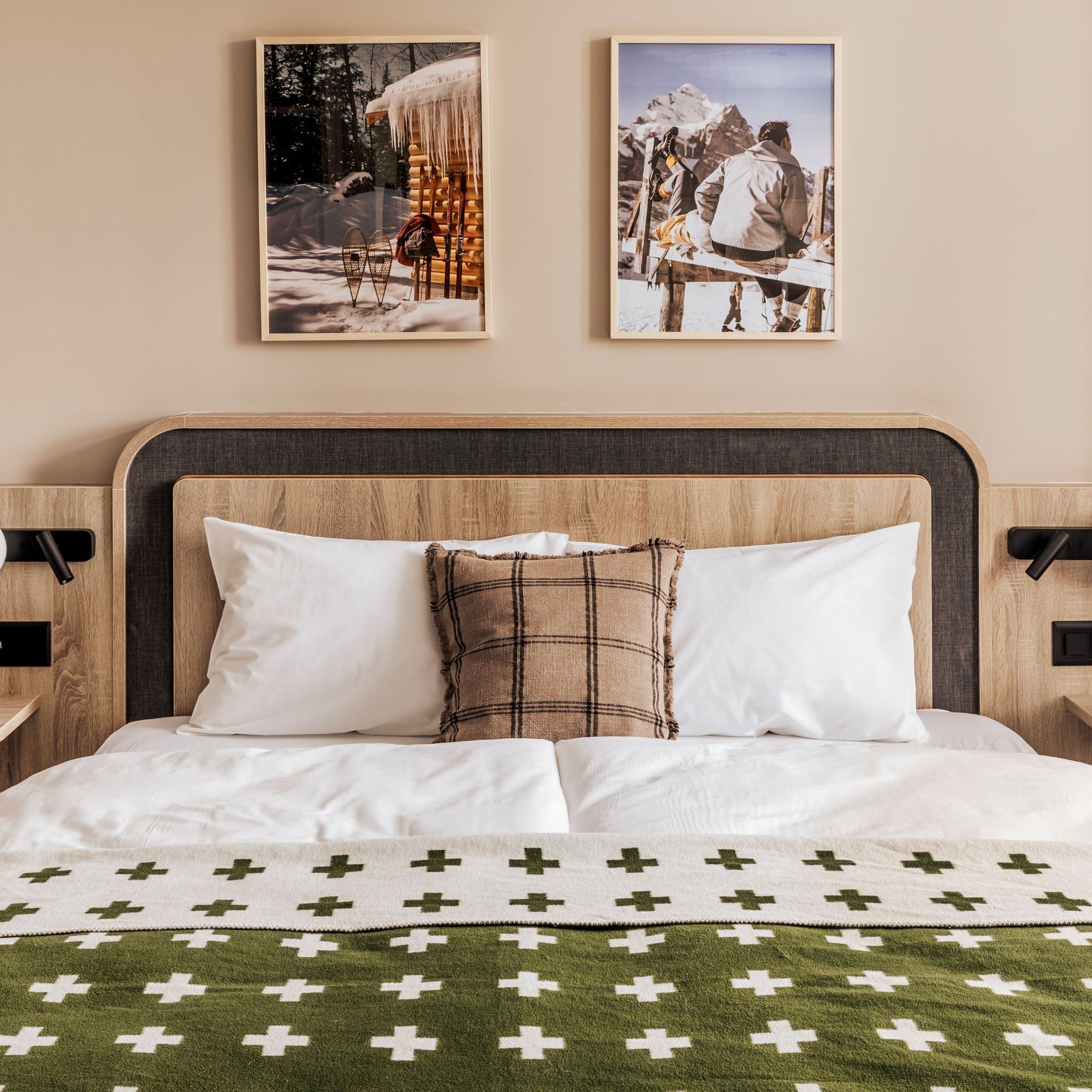 Un lit avec une literie blanche, un coussin à carreaux et une couverture à motifs verts et blancs. Deux tableaux encadrés sont accrochés au mur au-dessus du lit.