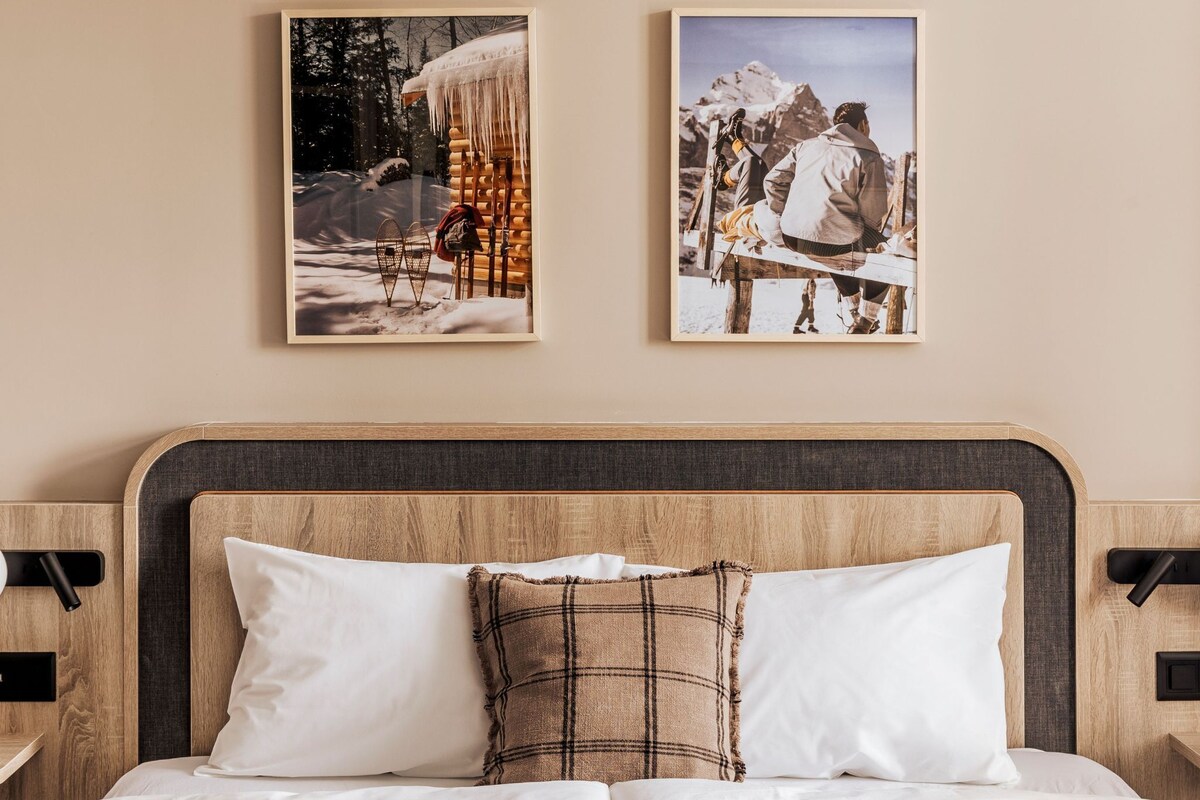 Ein Bett mit weißer Bettwäsche, einem karierten Kopfkissen und einer grün-weißen Tagesdecke. Darüber an der Wand hängen zwei gerahmte Bilder.