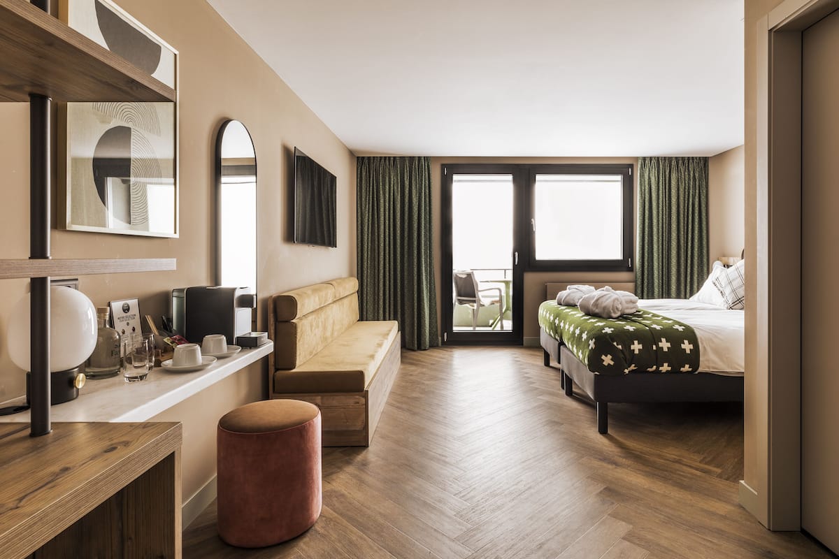 A modern hotel room with a wooden floor, a sofa, a bed with a green blanket, a desk with a lamp, and a large window with curtains.