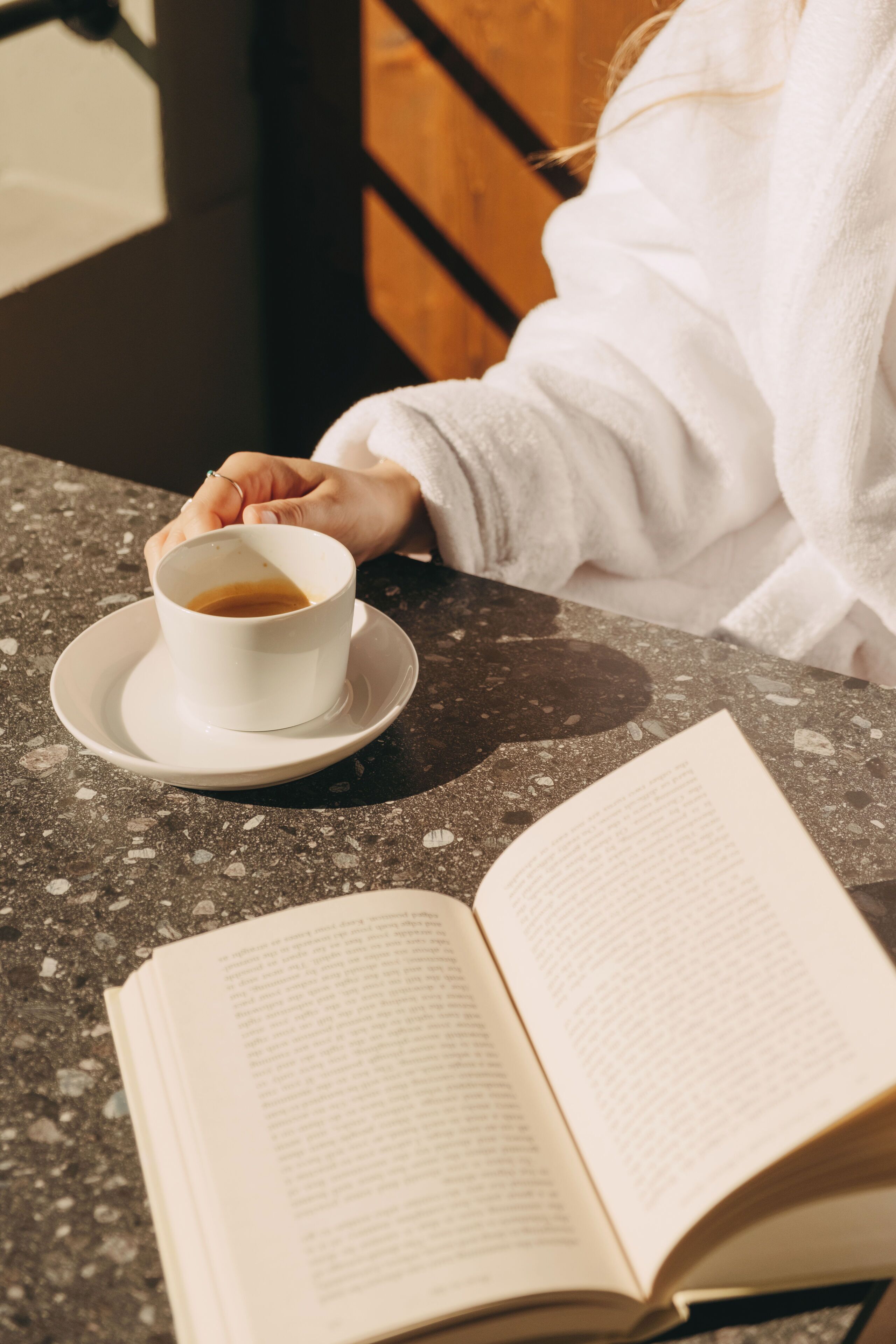 Una persona amb una bata blanca asseguda a taula, amb una tassa de cafè i un llibre obert.
