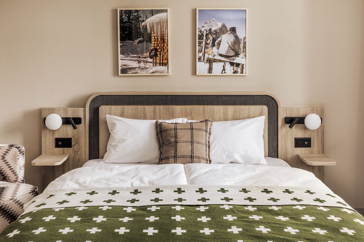 A neatly made bed with a green and white cross-patterned blanket, two pillows, and a wooden headboard. There are two wall-mounted nightstands with lamps, and two framed pictures above the bed.