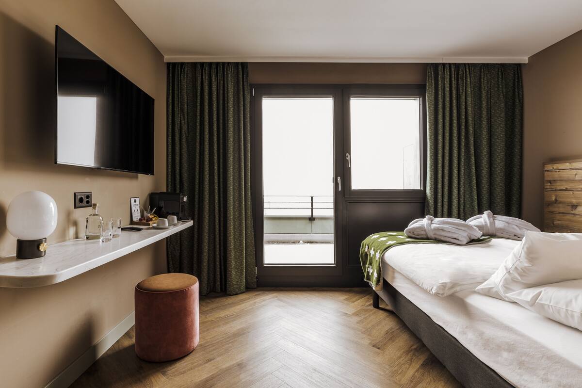 A hotel room with a bed, a desk with a flat-screen TV, a small round ottoman, and a view of the outdoors through a sliding door.