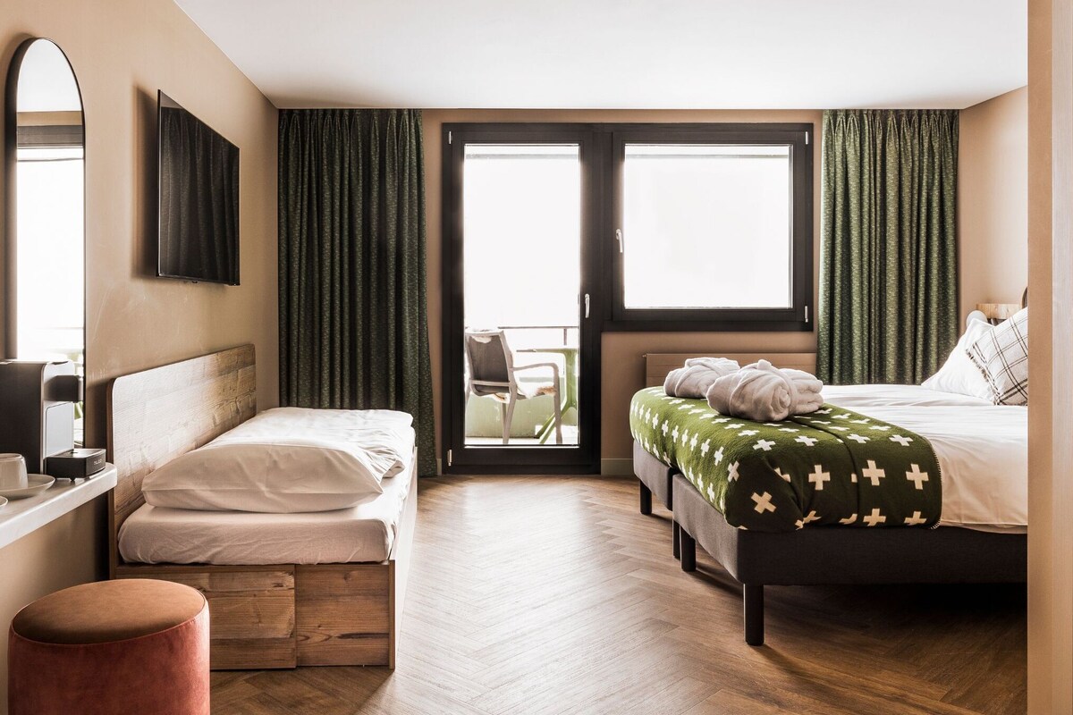 Hotelkamer met twee bedden, een houten vloer, een bedspread met groene en witte patronen, een rond poefje, een bureau met stoel, een flatscreen-tv en een glazen schuifdeur naar een balkon.