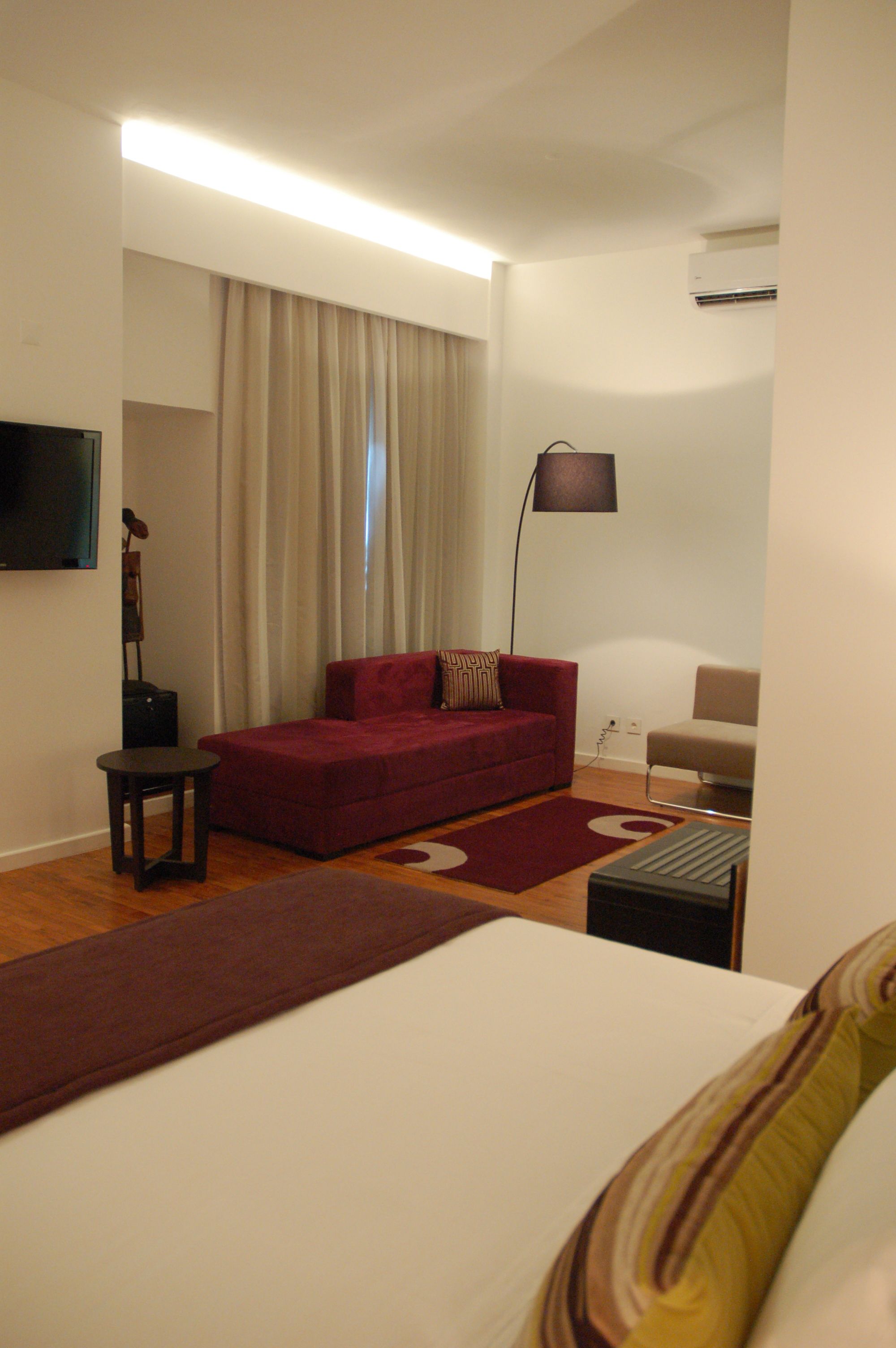 A hotel room with a bed, a red sofa, a small table, a lamp, and a TV on the wall.