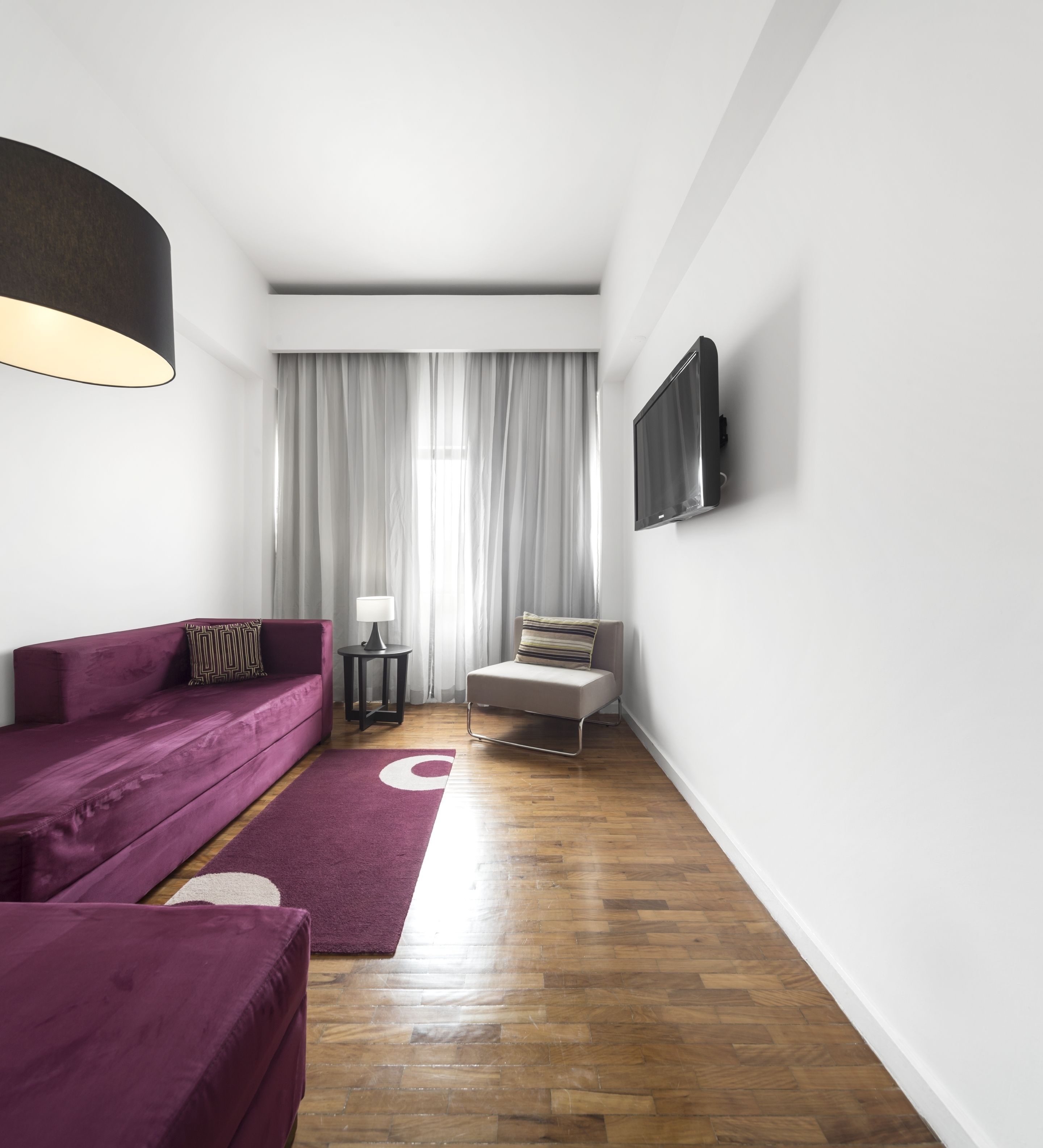 A modern living room with a purple sofa, a small table with a lamp, and a TV mounted on the wall.