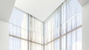 A dining area with a table set for two, large windows with sheer curtains, and a view of buildings outside.