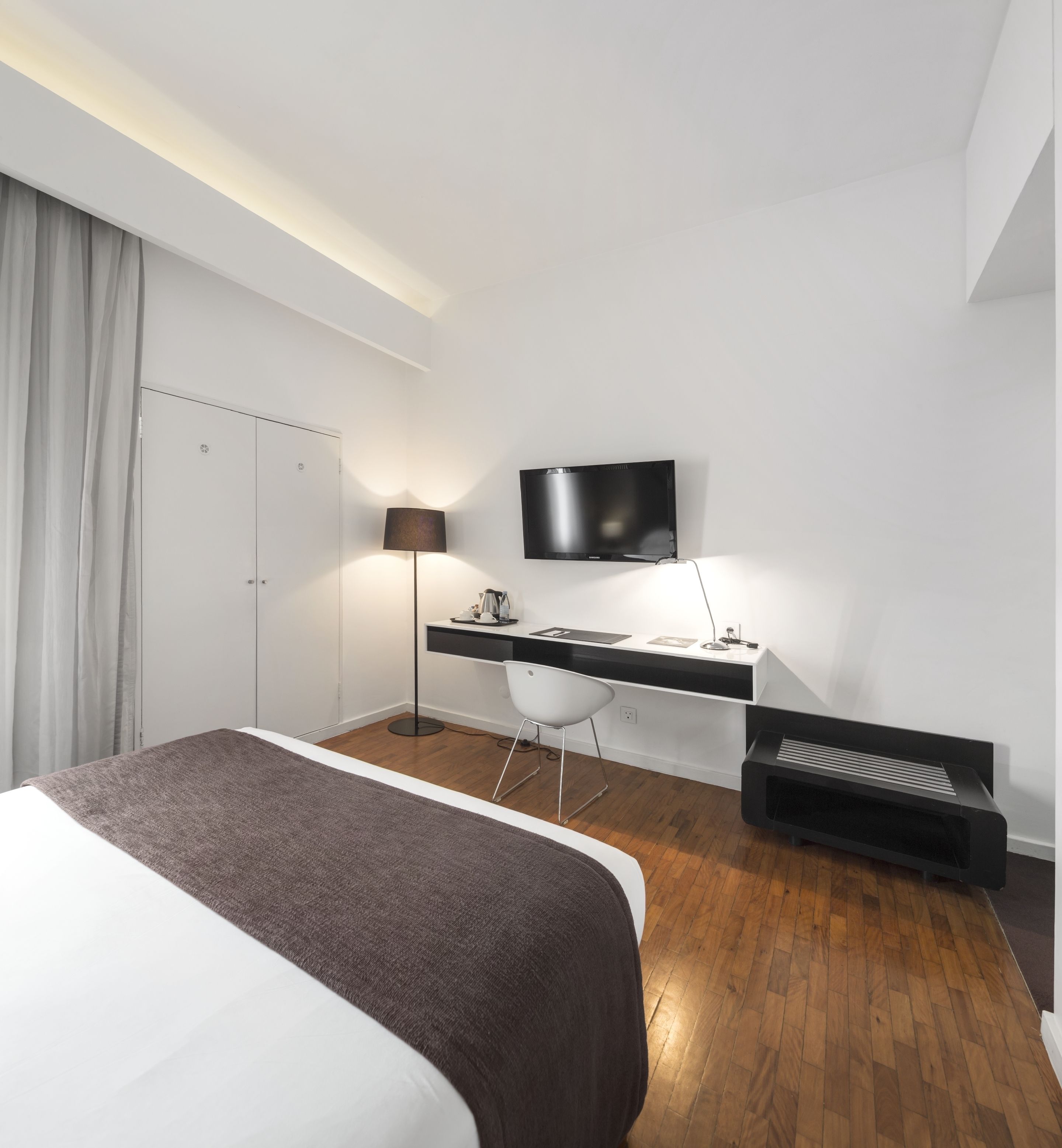 A modern hotel room with a bed, desk, chair, TV, and a small heater.