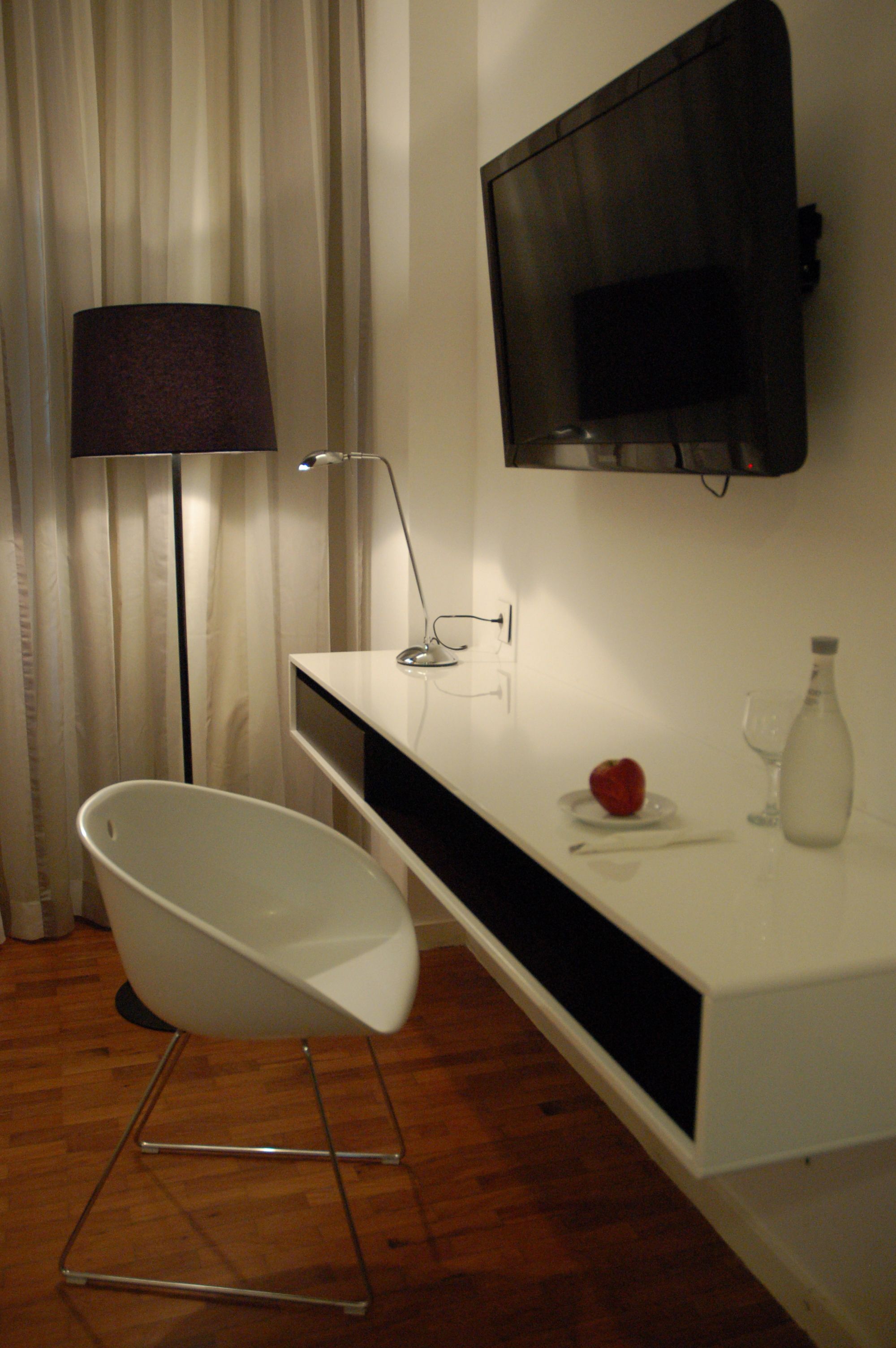 A modern bathroom with a white bathtub, a mounted TV, a lamp, and a small table with a bottle and a glass.