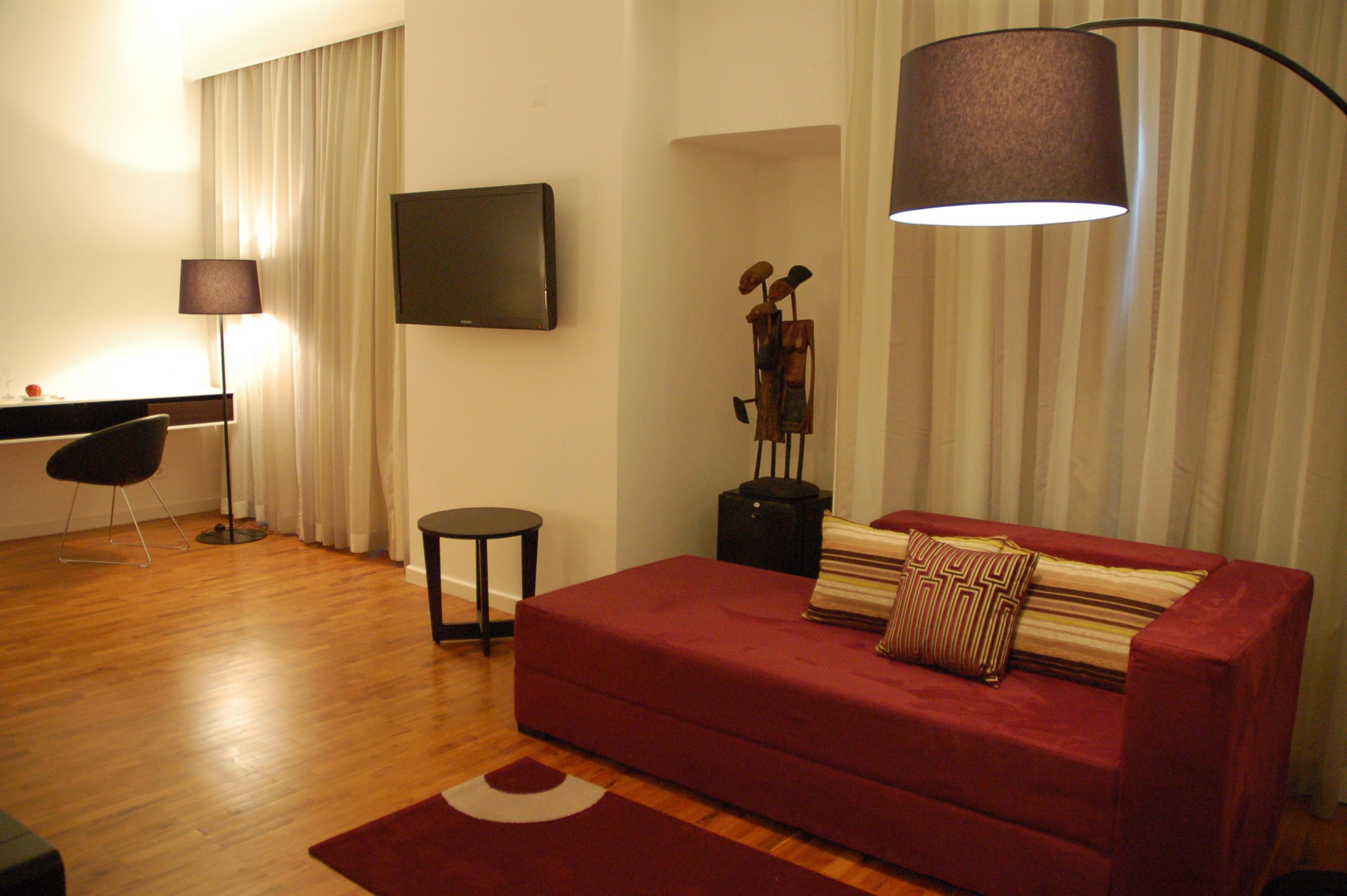 A modern living room with a red sofa, a small round table, a floor lamp, and a flat-screen TV mounted on the wall.