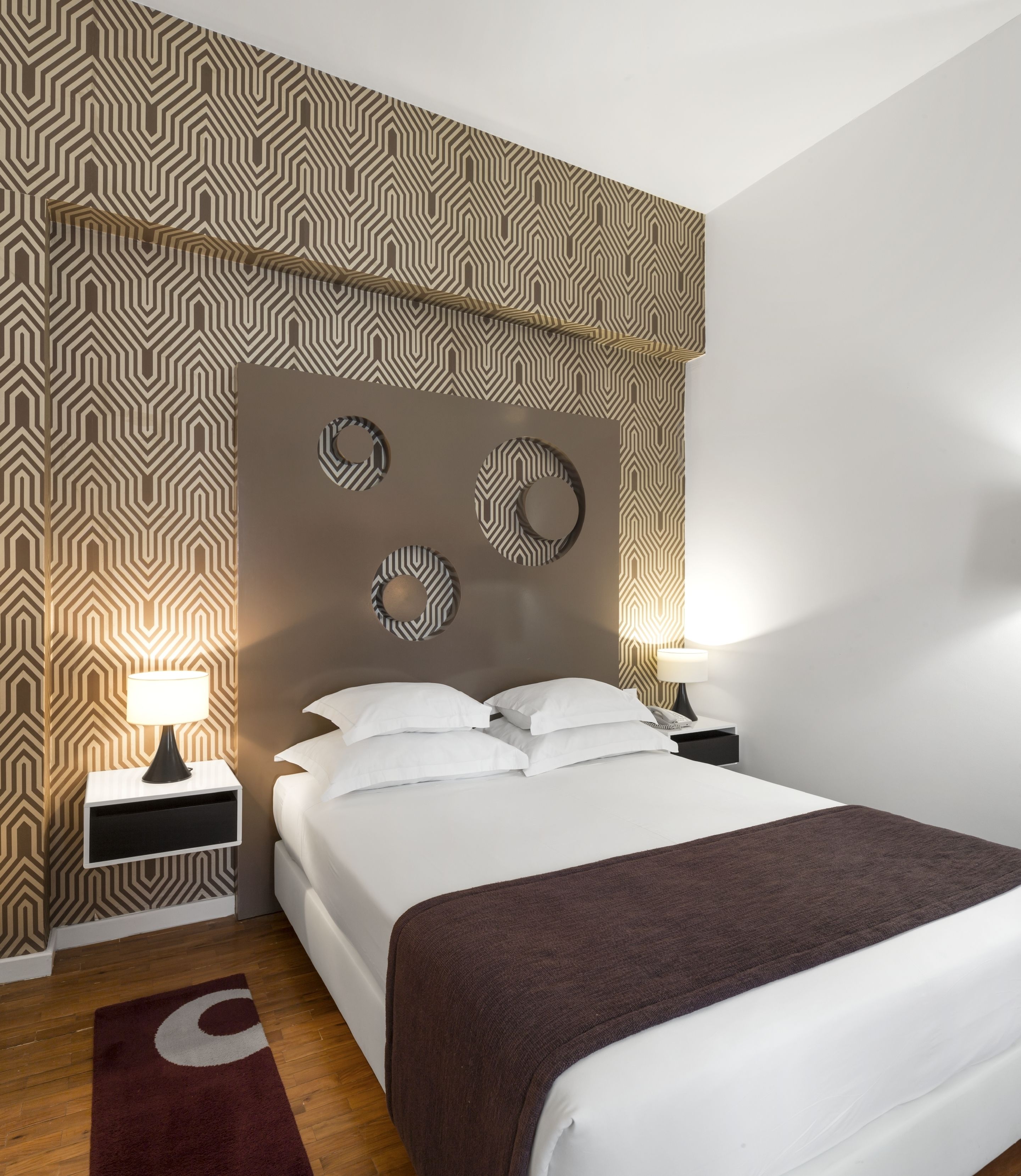 A modern bedroom with a patterned wallpaper, a large bed with white linens, two bedside tables with lamps, and a circular wall decoration.