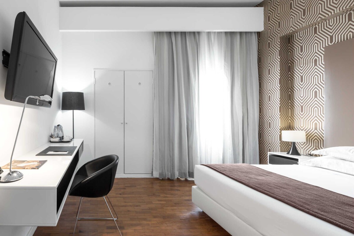 A hotel room with a bed, a desk with a chair, a television, and a wardrobe.
