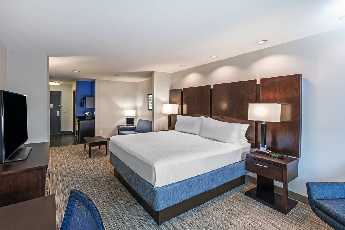 A hotel room with a large bed, a desk, a chair, a TV, and a nightstand with a lamp.