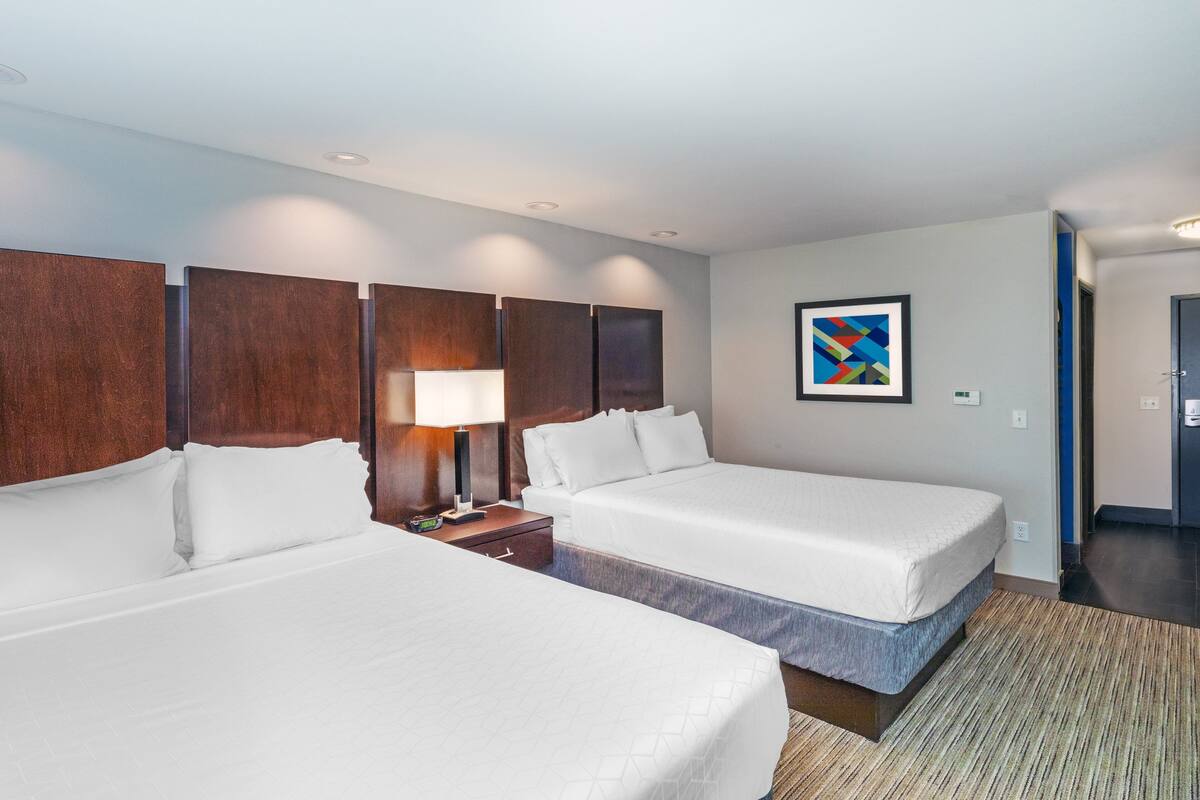A hotel room with two beds, a nightstand with a lamp, a wall-mounted artwork, and a door leading to another room.