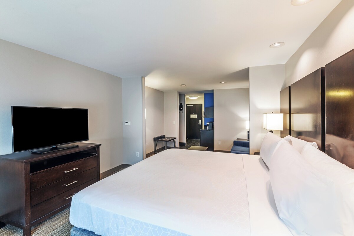A hotel room with a large bed, a flat-screen TV on a wooden dresser, a desk with a chair, and a mirror on the wall.