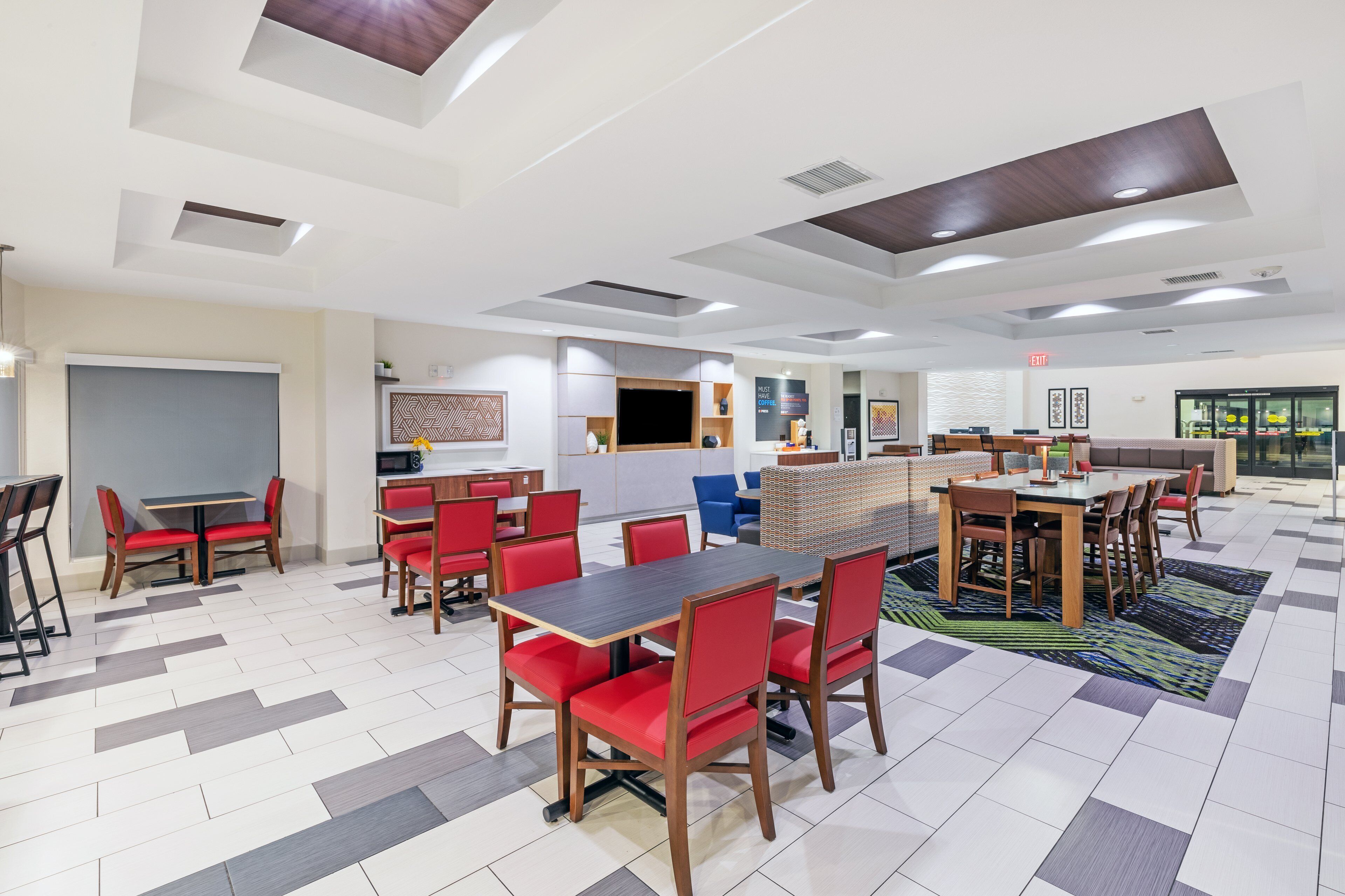 A spacious hotel dining area with multiple tables and chairs, a flat-screen TV, and a modern ceiling design.