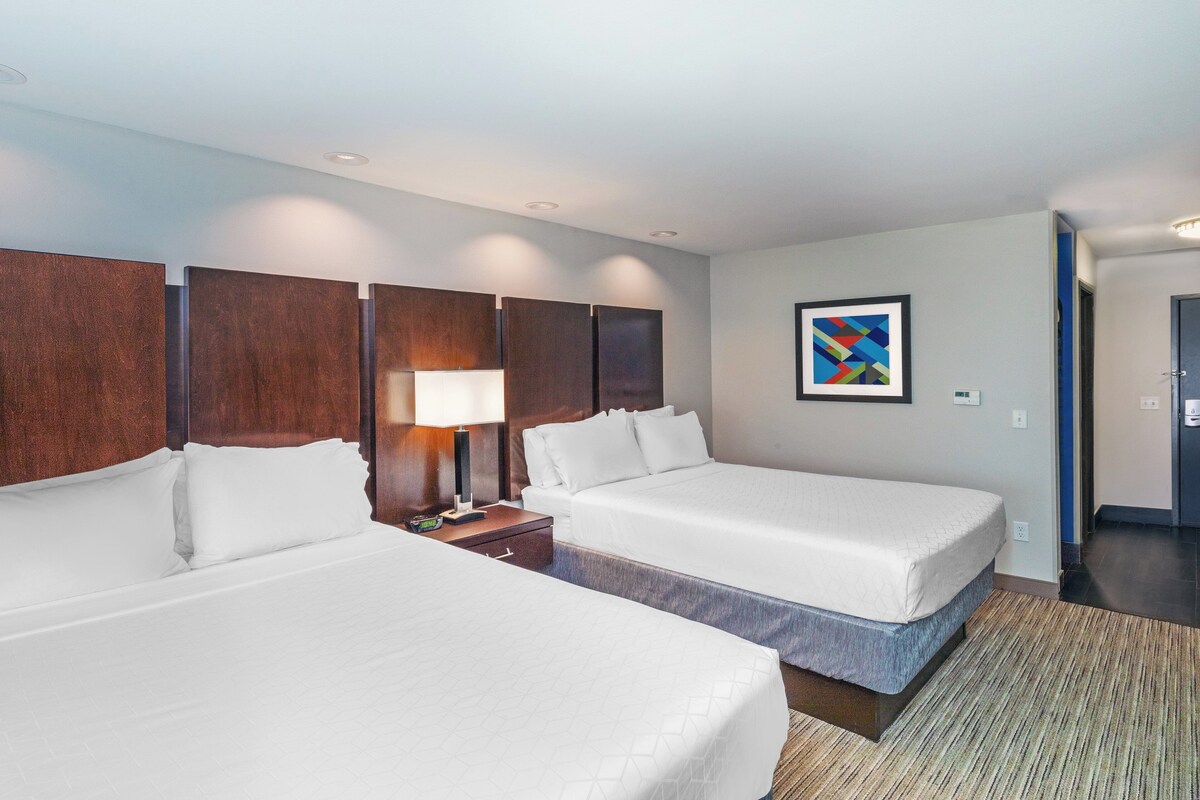 A hotel room with two beds, a nightstand, a lamp, and a painting on the wall.