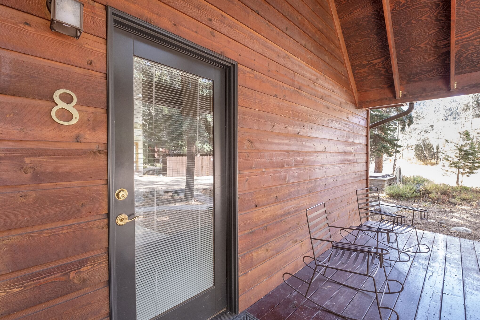 A wooden exterior with a door marked with the number 8, a covered patio with metal chairs, and a view of trees and a building in the background.