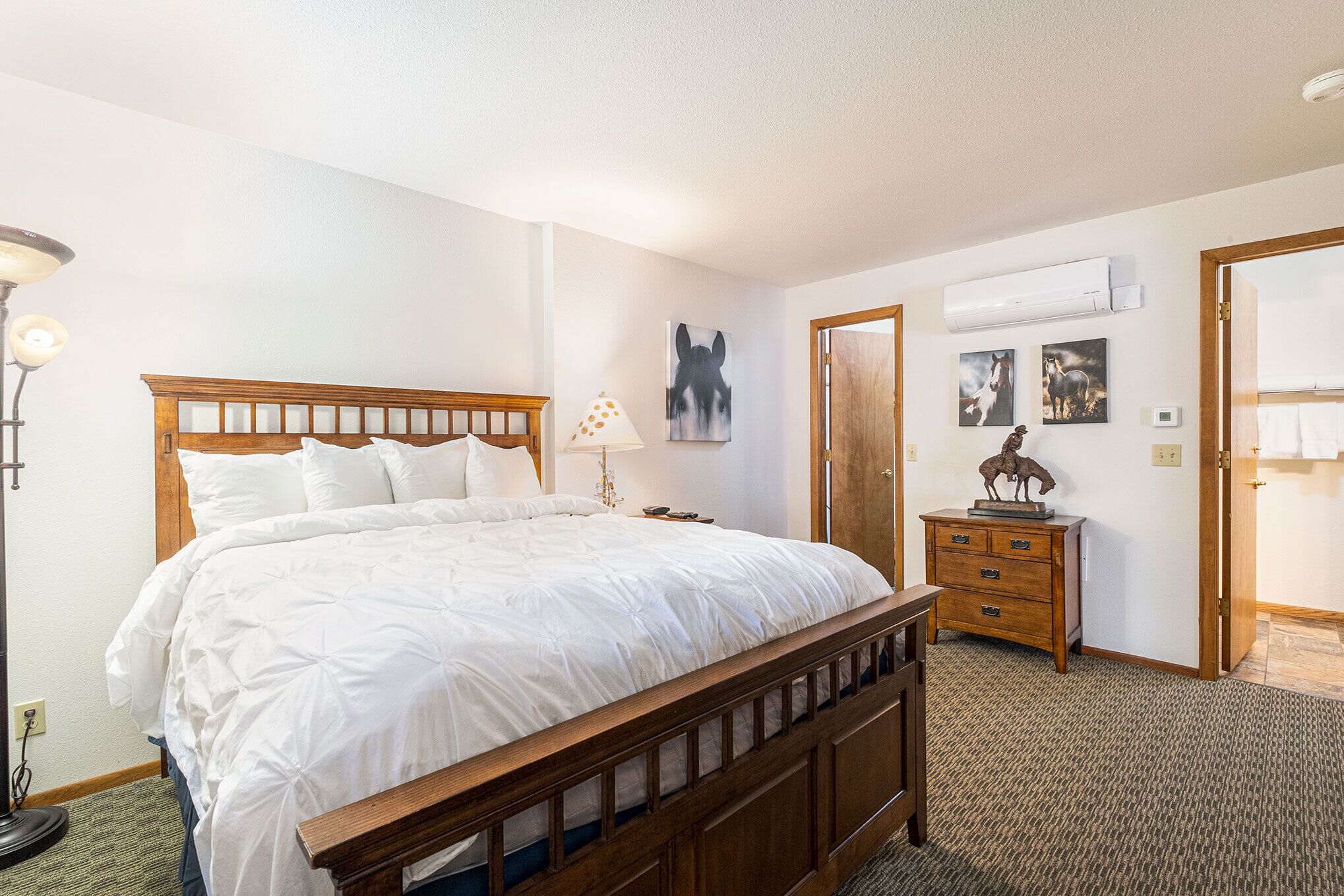 Een slaapkamer met een houten bed, een bedlampje, een dressoir en een deur die naar een andere kamer leidt.
