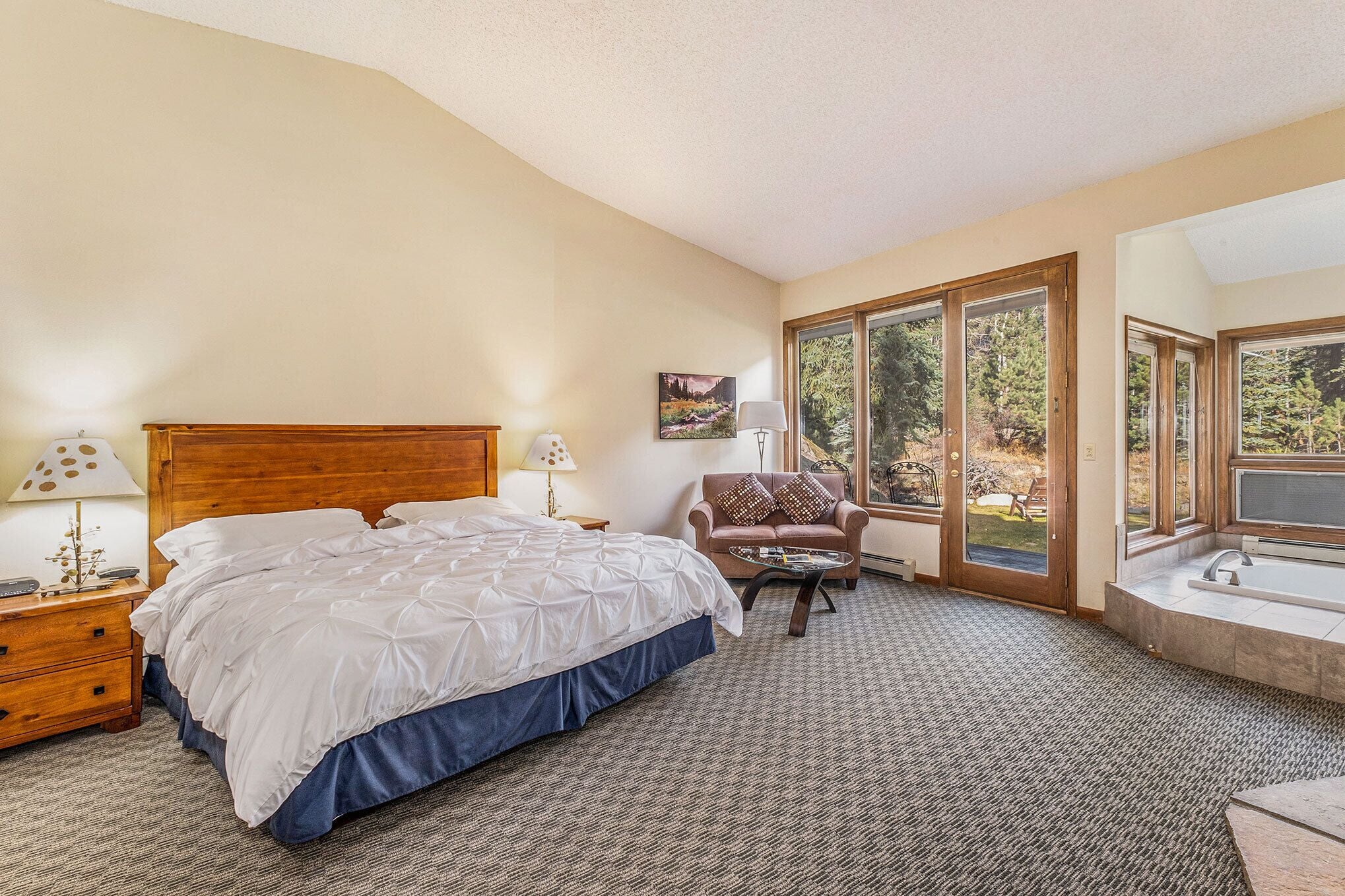 A bedroom with a large bed, a wooden headboard, a nightstand with a lamp, a chair with cushions, and a view of the outdoors through large windows.