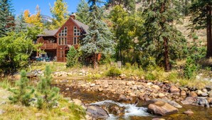 A wooden lodge with a stone foundation, surrounded by a natural landscape with a stream and trees. - Boulder Brook on Fall River (Estes Park)